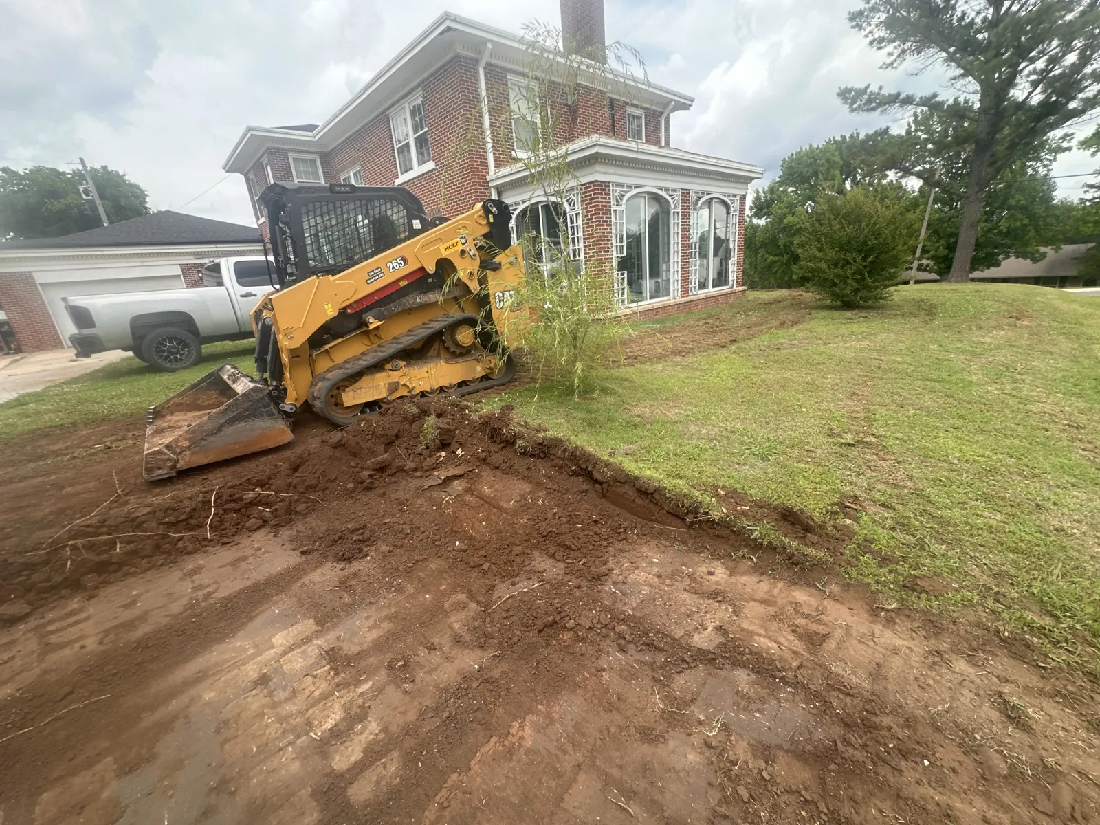 Tracked skid steer grading the dirt beside a brick house and lawn
